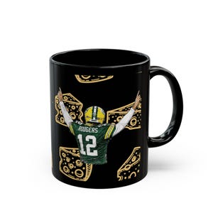 May include: A black ceramic mug featuring a graphic of a football player wearing a green and yellow jersey with the name RODGERS and the number 12 on the back, with arms raised, surrounded by yellow Swiss cheese wedge illustrations.