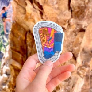 May include: A sticker in the shape of a carabiner with the words "Climb On" in a colorful design. The sticker features a blue and purple mountain scene with a climber and a blue device. The background is a rock climbing wall.