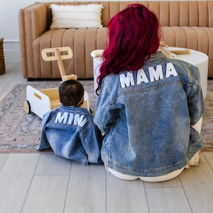 May include: Two denim jackets, one for an adult and one for a child, with the words "MAMA" and "MINI" in white lettering on the back. The adult has bright red hair. A wooden toy wagon is in the background.