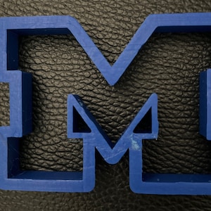 May include: A blue plastic cookie cutter in the shape of the letter "M". The cutter has a simple, blocky design with sharp angles. The cutter is made of a solid, opaque blue plastic.