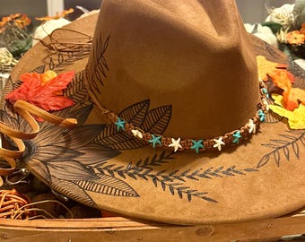 Pode incluir: Um chapéu de cowboy de feltro castanho com uma faixa decorativa de estrelas brancas e contas turquesas. A aba é adornada com um design floral e detalhes de folhas. O chapéu é exibido numa bandeja de madeira com folhagem de outono.