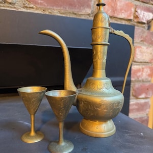 Puede incluir: Un juego de té de latón vintage, que incluye una jarra alta y ornamentada con un pico largo y un asa, y dos copas pequeñas. La jarra y las copas presentan diseños florales grabados. El juego se exhibe sobre una superficie oscura contra un fondo de ladrillos.
