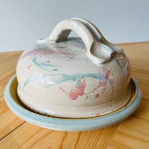 Può includere: Un porta burro in ceramica con manico e coperchio. Il piatto è decorato con una glassa bianca e spruzzi rosa e verdi.