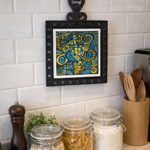 May include: A black framed decorative tile with a colorful abstract design in shades of blue, yellow, and green hangs on a white tiled wall. Below, glass jars filled with oats, pasta, and flour sit on a countertop, alongside a wooden utensil holder.