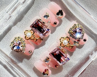May include: Pink and clear press-on nails with gold accents and pink, clear, and purple rhinestones. The nails are in a clear plastic case.