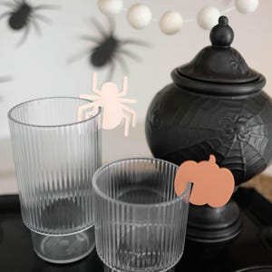 May include: Two clear glass cups with a pink spider and an orange pumpkin cutout on the rim. The cups are on a black tray with a black ceramic cauldron with a spiderweb design in the background.