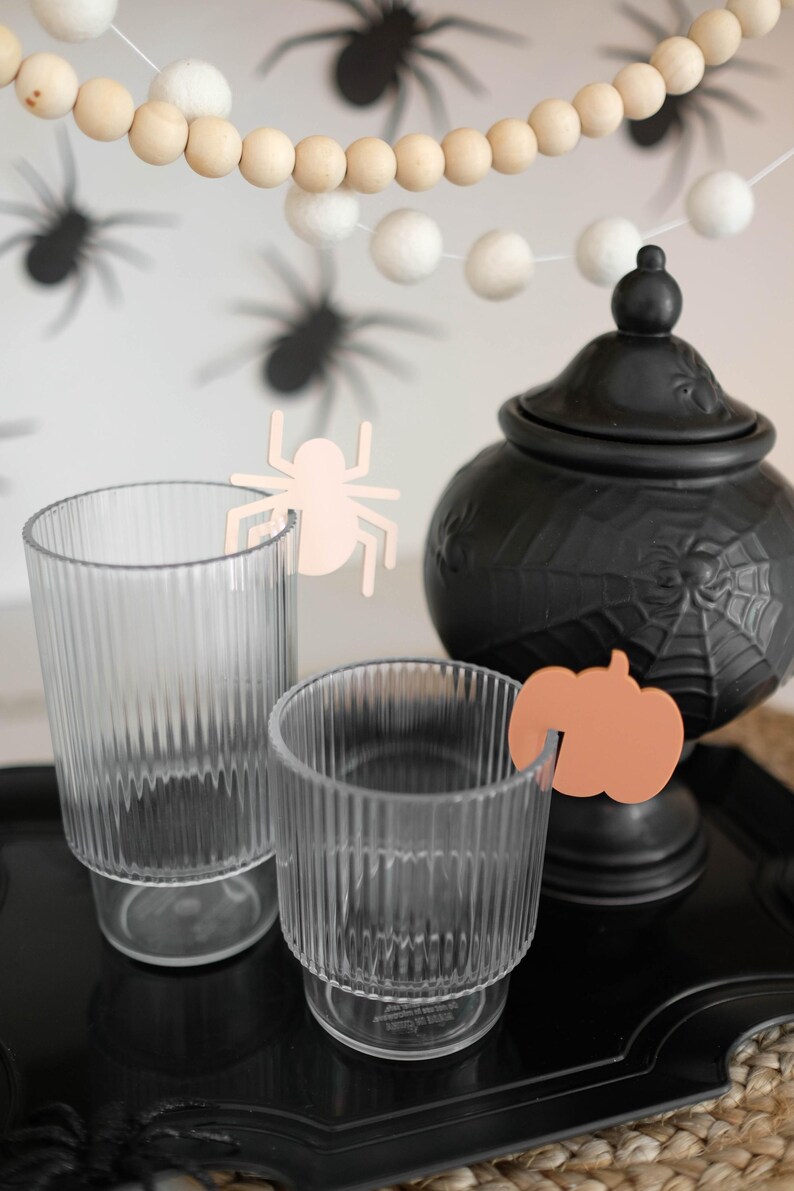 May include: Two clear glass cups with a pink spider and an orange pumpkin cutout on the rim. The cups are on a black tray with a black ceramic cauldron with a spiderweb design in the background.