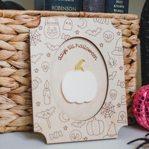 May include: Wooden Halloween countdown plaque featuring a white pumpkin and the words "days till halloween". The frame is decorated with Halloween-themed illustrations. The plaque is displayed in front of a woven basket and books.