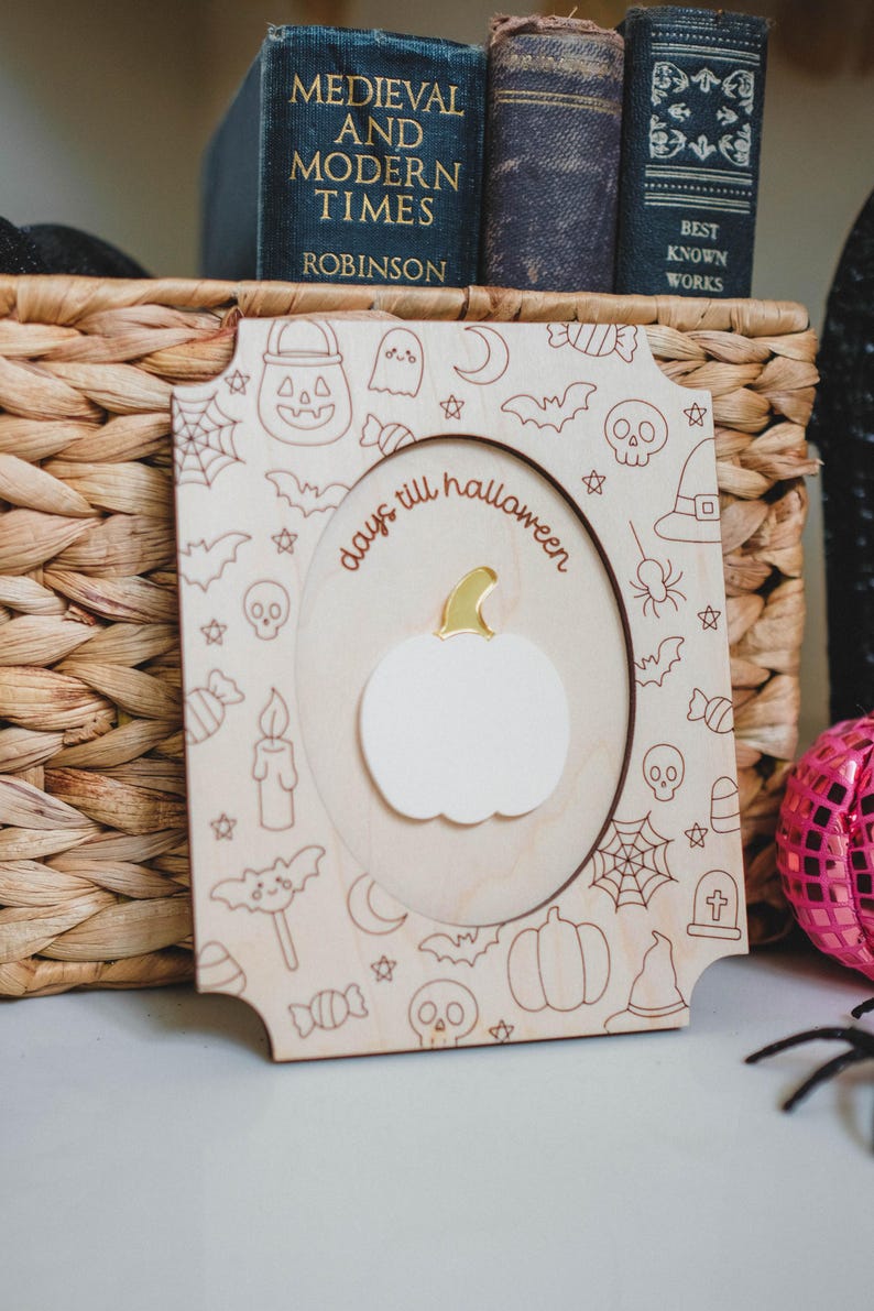 May include: Wooden Halloween countdown plaque featuring a white pumpkin and the words "days till halloween". The frame is decorated with Halloween-themed illustrations. The plaque is displayed in front of a woven basket and books.