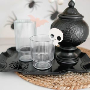 May include: A black tray with two clear glasses and a black ceramic jar with a spiderweb design. A white skull-shaped ornament is on the tray. The tray is on a woven brown placemat.