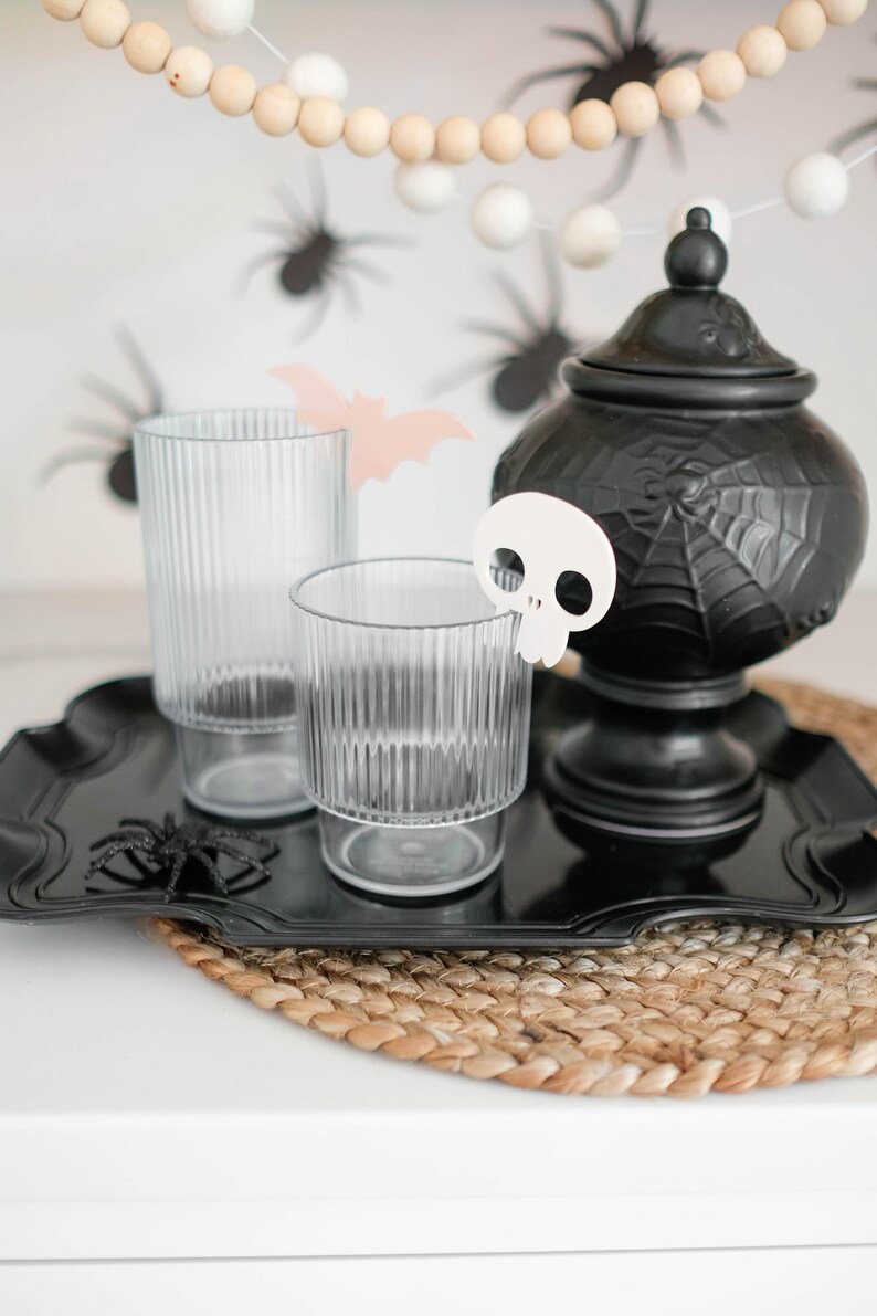 May include: A black tray with two clear glasses and a black ceramic jar with a spiderweb design. A white skull-shaped ornament is on the tray. The tray is on a woven brown placemat.