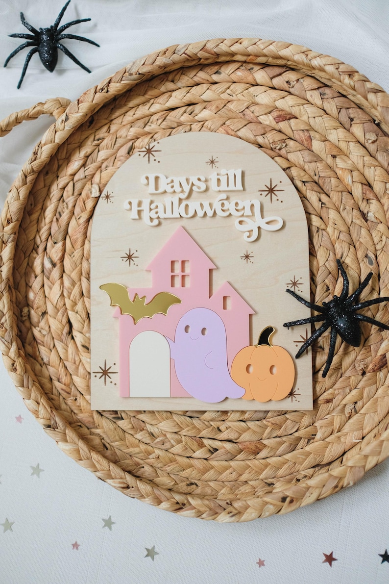 May include: Wooden Halloween countdown sign with a pink house, ghost, and pumpkin. The sign reads "Days till Halloween" in white lettering. Includes a gold bat and star accents. Two black glitter spiders are placed on the woven tray.