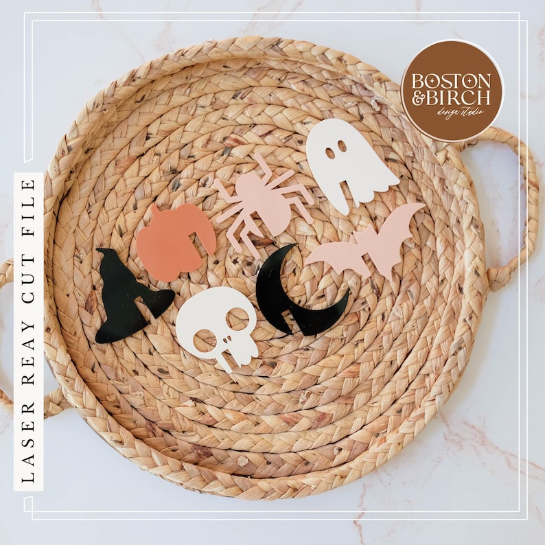 May include: A set of seven Halloween-themed laser cut shapes in various colors, including black, white, pink, and orange. The shapes include a ghost, a spider, a bat, a crescent moon, a skull, a witch's hat, and a pumpkin. The shapes are arranged on a woven straw mat.