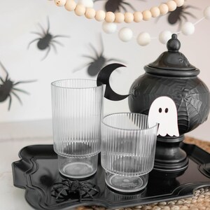 May include: A black tray with two clear glass cups, a black ceramic teapot with a ghost and spiderweb design, and a black spider on the tray. The tray is on a woven placemat. A white and wood bead garland with white pom-poms hangs above the tray.
