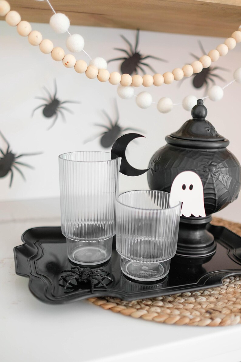May include: A black tray with two clear glass cups, a black ceramic teapot with a ghost and spiderweb design, and a black spider on the tray. The tray is on a woven placemat. A white and wood bead garland with white pom-poms hangs above the tray.