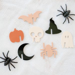 May include: A set of nine Halloween-themed paper cutouts, including a bat, a ghost, a witch's hat, a skull, a pumpkin, a spider, a crescent moon, and two spiders. The cutouts are on a white background.