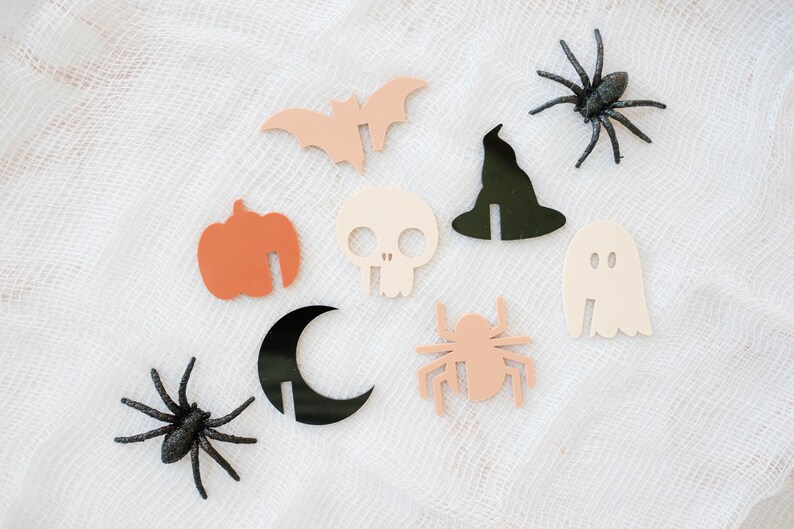 May include: A set of nine Halloween-themed paper cutouts, including a bat, a ghost, a witch's hat, a skull, a pumpkin, a spider, a crescent moon, and two spiders. The cutouts are on a white background.