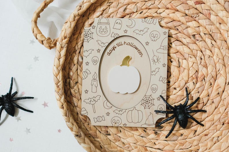 May include: A wooden Halloween countdown plaque with a white pumpkin and gold stem. The plaque is decorated with Halloween-themed doodles and the words "days till halloween". Two black plastic spiders are on the sides.
