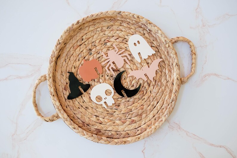 May include: A wicker tray with a variety of Halloween-themed decorations, including a ghost, a bat, a spider, a witch's hat, a skull, and a crescent moon.
