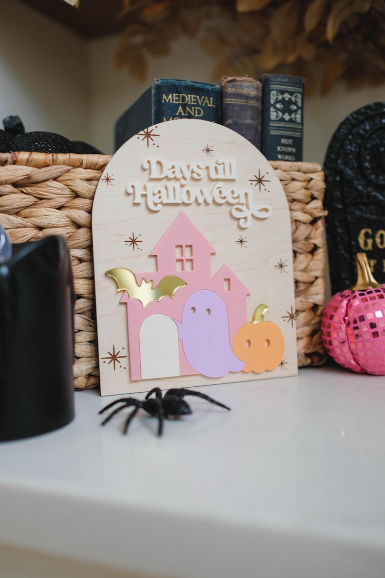 May include: Wooden Halloween sign with a pink house, ghost, and pumpkin. The sign reads "Days till Halloween" and features a gold bat. A black spider sits in front of the sign.