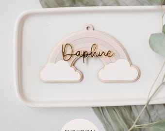 Könnte beinhalten: Ein lasergeschnittener Holzregenbogen mit dem goldenen Namen "Daphne" darauf. Der Regenbogen ist in verschiedenen Rosatönen und Weiß geschichtet. Der Regenbogen befindet sich auf einem weißen Teller mit einer grünen Pflanze im Hintergrund.