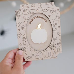 May include: Wooden Halloween countdown plaque with a pumpkin cutout and the words "days till halloween". The frame is decorated with engraved Halloween-themed icons like bats, ghosts, and candy. The pumpkin has a gold stem.
