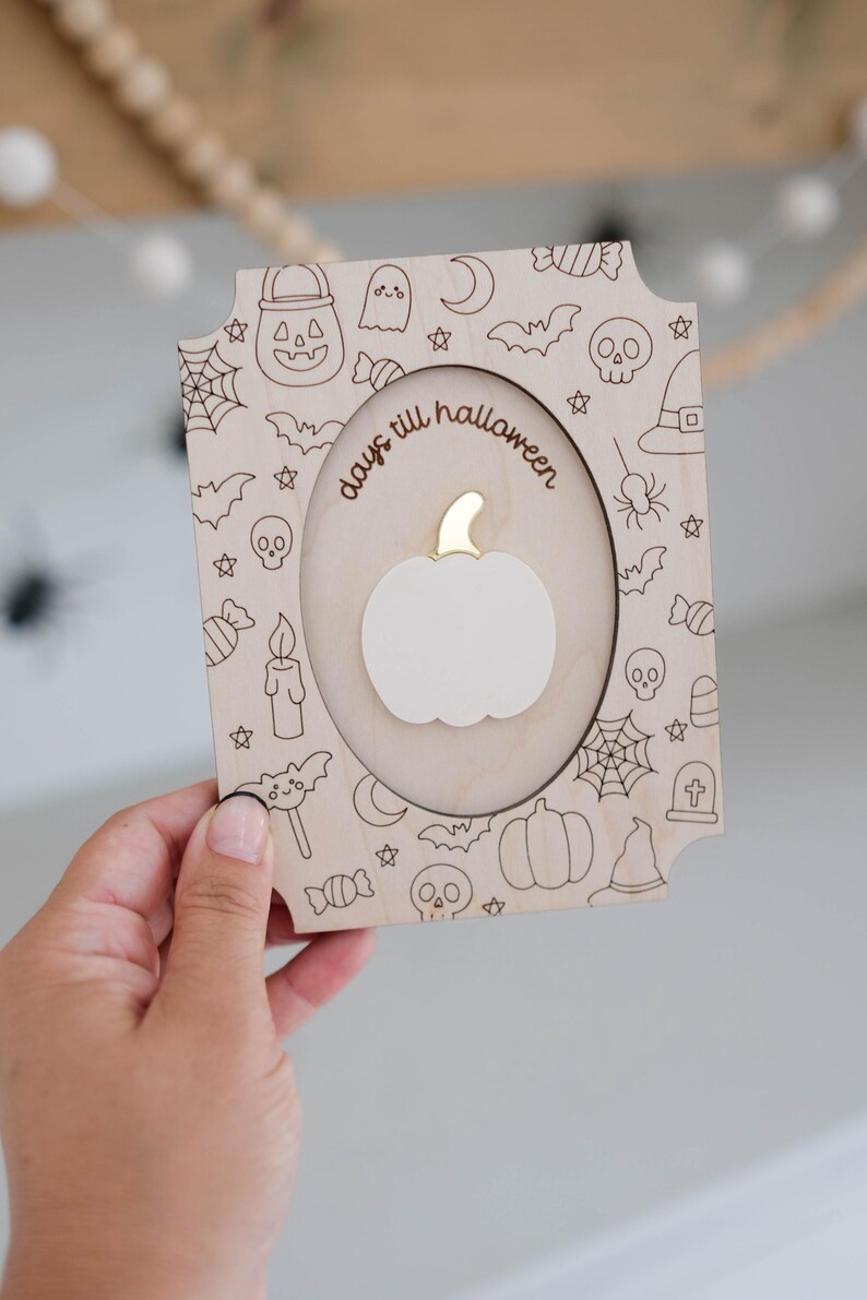 May include: Wooden Halloween countdown plaque with a pumpkin cutout and the words "days till halloween". The frame is decorated with engraved Halloween-themed icons like bats, ghosts, and candy. The pumpkin has a gold stem.