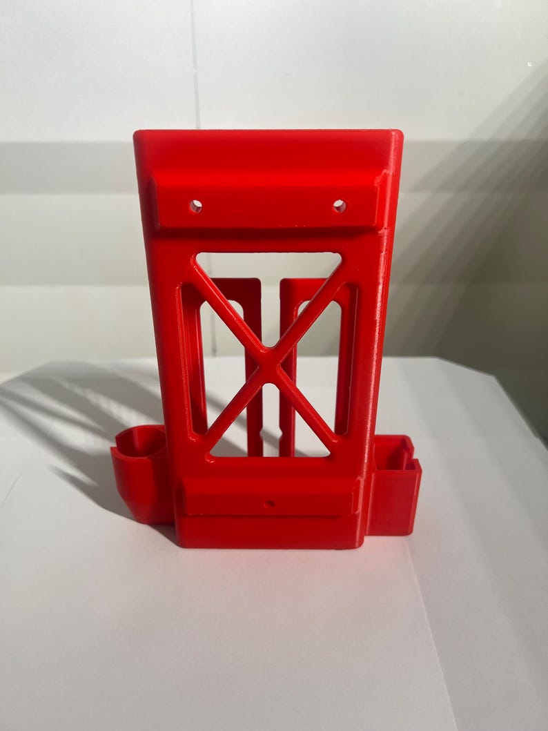 K&ouml;nnte beinhalten: Ein leuchtend rotes, 3D-gedrucktes Objekt mit einem rechteckigen Rahmen und einem offenen, geometrischen Design. Es hat zwei kleine Seitenf&auml;cher und eine zentrale, kreuzf&ouml;rmige St&uuml;tzstruktur. Oben sind zwei L&ouml;cher sichtbar.