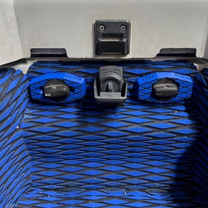 K&ouml;nnte beinhalten: Nahaufnahme eines Jet-Ski-Fachs mit blau-schwarzer, rautenf&ouml;rmiger Polsterung. Merkmale sind ein zentraler Riegel mit der Aufschrift "LOCK OPEN" und zwei blaue Bedienkn&ouml;pfe mit der Aufschrift "CHOKE" und "ON". Das Fach ist wei&szlig;.