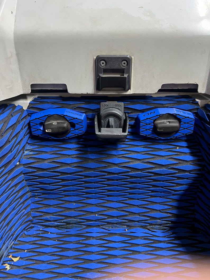 K&ouml;nnte beinhalten: Nahaufnahme eines Jet-Ski-Fachs mit blau-schwarzer, rautenf&ouml;rmiger Polsterung. Merkmale sind ein zentraler Riegel mit der Aufschrift "LOCK OPEN" und zwei blaue Bedienkn&ouml;pfe mit der Aufschrift "CHOKE" und "ON". Das Fach ist wei&szlig;.