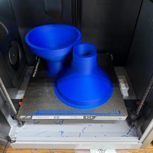 K&ouml;nnte beinhalten: Ein 3D-Drucker mit zwei gro&szlig;en, leuchtend blauen Kunststoffobjekten, die gerade gedruckt werden. Die Objekte sind trichterf&ouml;rmig, wobei eines breiter ist als das andere. Der Drucker ist in einem grau-schwarzen Geh&auml;use eingeschlossen.