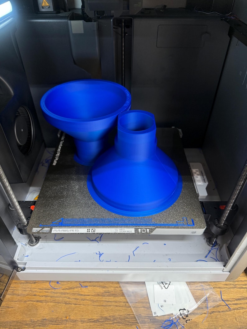 K&ouml;nnte beinhalten: Ein 3D-Drucker mit zwei gro&szlig;en, leuchtend blauen Kunststoffobjekten, die gerade gedruckt werden. Die Objekte sind trichterf&ouml;rmig, wobei eines breiter ist als das andere. Der Drucker ist in einem grau-schwarzen Geh&auml;use eingeschlossen.