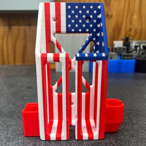 K&ouml;nnte beinhalten: Ein 3D-gedruckter Halter mit patriotischem Design. Der obere Bereich zeigt ein blaues Feld mit wei&szlig;en Sternen, w&auml;hrend der Hauptk&ouml;rper rote und wei&szlig;e Streifen aufweist. Zwei rote Seitenf&auml;cher sind enthalten.