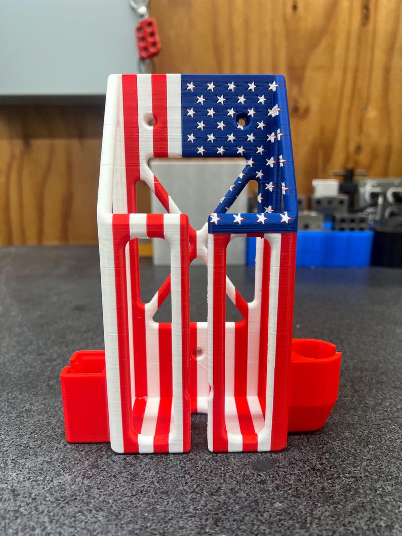 K&ouml;nnte beinhalten: Ein 3D-gedruckter Halter mit patriotischem Design. Der obere Bereich zeigt ein blaues Feld mit wei&szlig;en Sternen, w&auml;hrend der Hauptk&ouml;rper rote und wei&szlig;e Streifen aufweist. Zwei rote Seitenf&auml;cher sind enthalten.