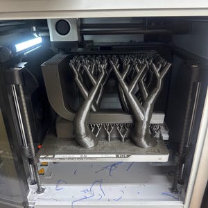 K&ouml;nnte beinhalten: Ein 3D-Drucker in Betrieb, der eine komplexe, baumartige Struktur erstellt. Das Objekt ist grau und scheint aus Kunststoff zu bestehen. Der Drucker ist in einem wei&szlig;en und grauen Geh&auml;use untergebracht, mit sichtbaren mechanischen Komponenten.