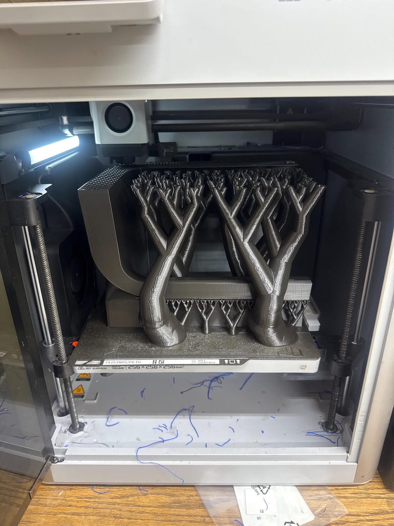 K&ouml;nnte beinhalten: Ein 3D-Drucker in Betrieb, der eine komplexe, baumartige Struktur erstellt. Das Objekt ist grau und scheint aus Kunststoff zu bestehen. Der Drucker ist in einem wei&szlig;en und grauen Geh&auml;use untergebracht, mit sichtbaren mechanischen Komponenten.