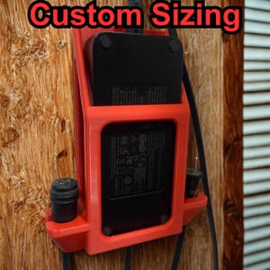 Puede incluir: Un soporte de carga rojo impreso en 3D con el texto "Custom Sizing" en rojo. El soporte contiene un dispositivo de carga negro y tiene un enchufe y un cable negros. El soporte está montado en una superficie de madera.