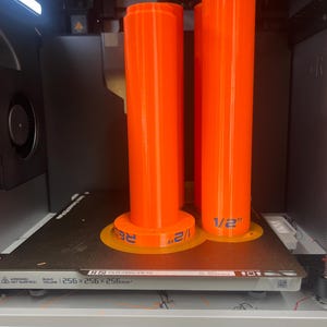 K&ouml;nnte beinhalten: Zwei leuchtend orangefarbene zylindrische Objekte mit kreisf&ouml;rmigen Basen befinden sich auf einem 3D-Druckerbett. Die Basen haben blauen Text mit der Aufschrift "1/2" und "RE". Das Druckerbett hat den Text "256x256x256mm".