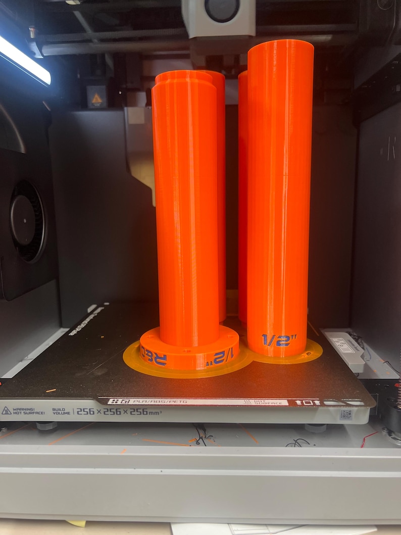 K&ouml;nnte beinhalten: Zwei leuchtend orangefarbene zylindrische Objekte mit kreisf&ouml;rmigen Basen befinden sich auf einem 3D-Druckerbett. Die Basen haben blauen Text mit der Aufschrift "1/2" und "RE". Das Druckerbett hat den Text "256x256x256mm".