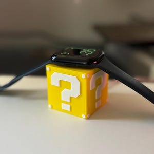 Puede incluir: Un Apple Watch Series 7 negro está sentado en un bloque amarillo con un signo de interrogación. La esfera del reloj muestra la hora.