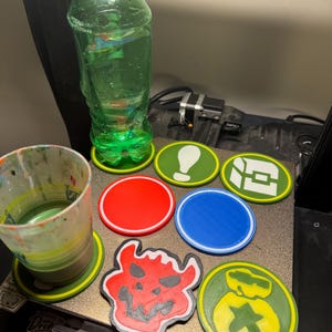Puede incluir: Un conjunto de siete posavasos impresos en 3D con varios diseños, que incluyen un círculo rojo, un círculo azul, un círculo verde con un signo de exclamación blanco, un círculo verde con un cofre blanco, una calavera de demonio roja y un círculo verde con una bolsa de dinero blanca y una estrella. Los posavasos están sobre una superficie negra.
