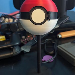 Könnte beinhalten: Eine rot-schwarz-weiße Pokéball-Replik, montiert auf einem schwarzen Ständer. Der Ball ist in rote und weiße Hälften mit einem schwarzen Band und einem weißen kreisförmigen Knopf unterteilt. Der Ständer hat einen kreisförmigen Sockel.