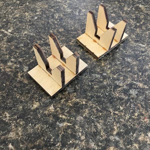 May include: Two small, unfinished wooden display stands. Each stand has a rectangular base with three upright sections. The wood has a natural, light brown color with darker edges. The stands are designed to hold items.