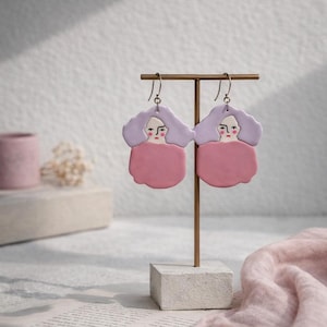 May include: A pair of handmade earrings featuring a unique design. Each earring showcases a face with lavender hair and a pink body. The earrings are displayed on a gold stand with a white base, set against a soft, neutral background.