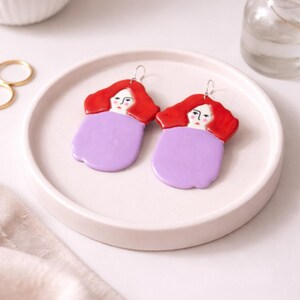 May include: A pair of handmade ceramic earrings. Each earring features a red-haired woman's face atop a lavender body. The earrings are on a white ceramic plate. The earrings are a unique and artistic accessory.