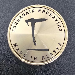 May include: A gold-colored coin with the text "TURNAGAIN ENGRAVING" around the top edge and "MADE IN ALASKA" along the bottom. A black graphic of a stylized "T" is in the center.