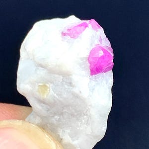 May include: A close-up of a white mineral specimen with bright pink crystal formations. The rough, textured surface of the stone contrasts with the sharp, angular crystals. The specimen is held against a dark background.