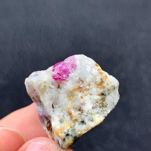 May include: A small, rough gemstone featuring a bright pink ruby crystal set within a matrix of white and tan minerals. The stone has a natural, unpolished look, highlighting its raw beauty. The background is a dark grey.