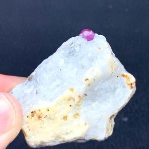 May include: A small, raw ruby gemstone sits atop a white and beige rock formation. The ruby is a deep pink color, and the rock has a rough, textured surface. The background is a dark blue.