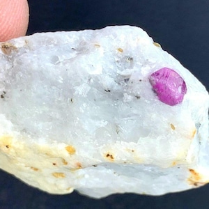 Peut inclure: Une pierre blanche brute avec un cristal de rubis rose vif incrusté. La pierre a une texture légèrement rugueuse avec de petits points sombres et une touche de coloration jaune-brun sur les bords. Le rubis est d'un rose profond et saturé.